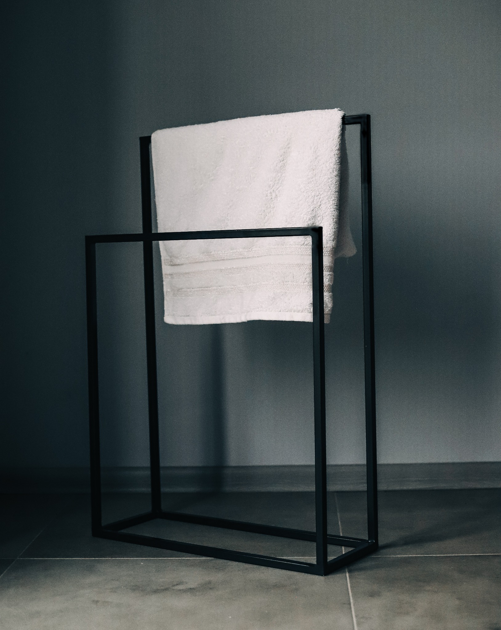 Minimalistyczny, czarny, metalowy stojak na ręczniki z białym, puszystym ręcznikiem na tle szarej ściany i podłogi z płytek.