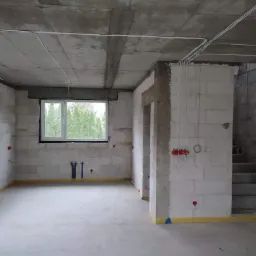Surowy stan wnętrza budynku w trakcie prac: białe ściany z betonu komórkowego, widoczne przewody elektryczne, puszki instalacyjne oraz przygotowane podejścia hydrauliczne. Okna z widokiem na zieleń...