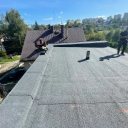 Nowe, czarne pokrycie dachu z papy termozgrzewalnej na tle zabudowy miejskiej i zieleni, dekarz stoi na dachu.