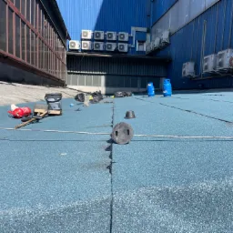 Prace na dachu przemysłowym z niebieską papą, widoczne narzędzia, butle gazowe, pracownik z palnikiem oraz liczne klimatyzatory na ścianie budynku.