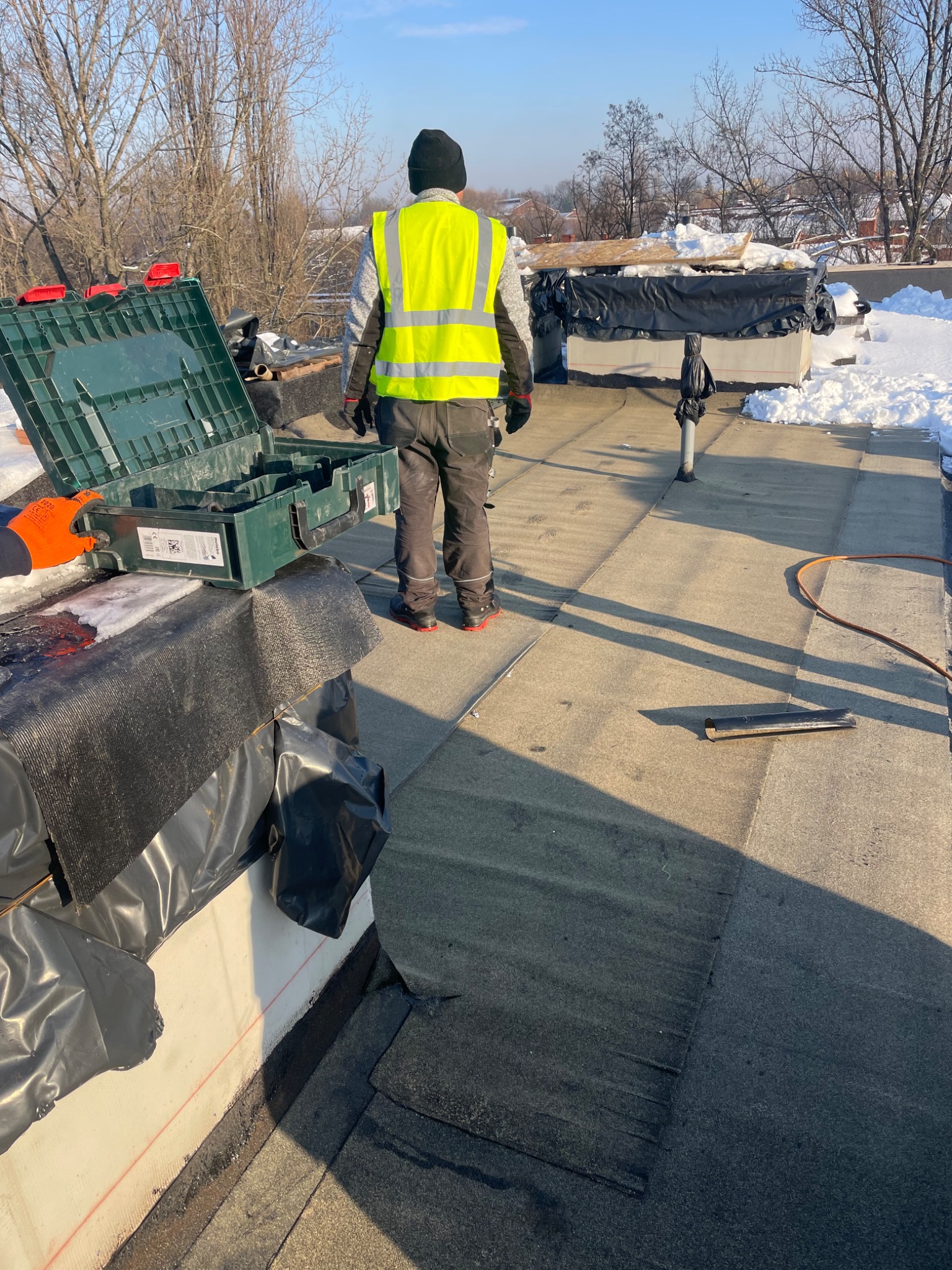Pracownik w kamizelce odblaskowej na płaskim dachu podczas prac dekarskich w zimowych warunkach, widoczna skrzynka z narzędziami, częściowo ułożona papa i śnieg w tle.