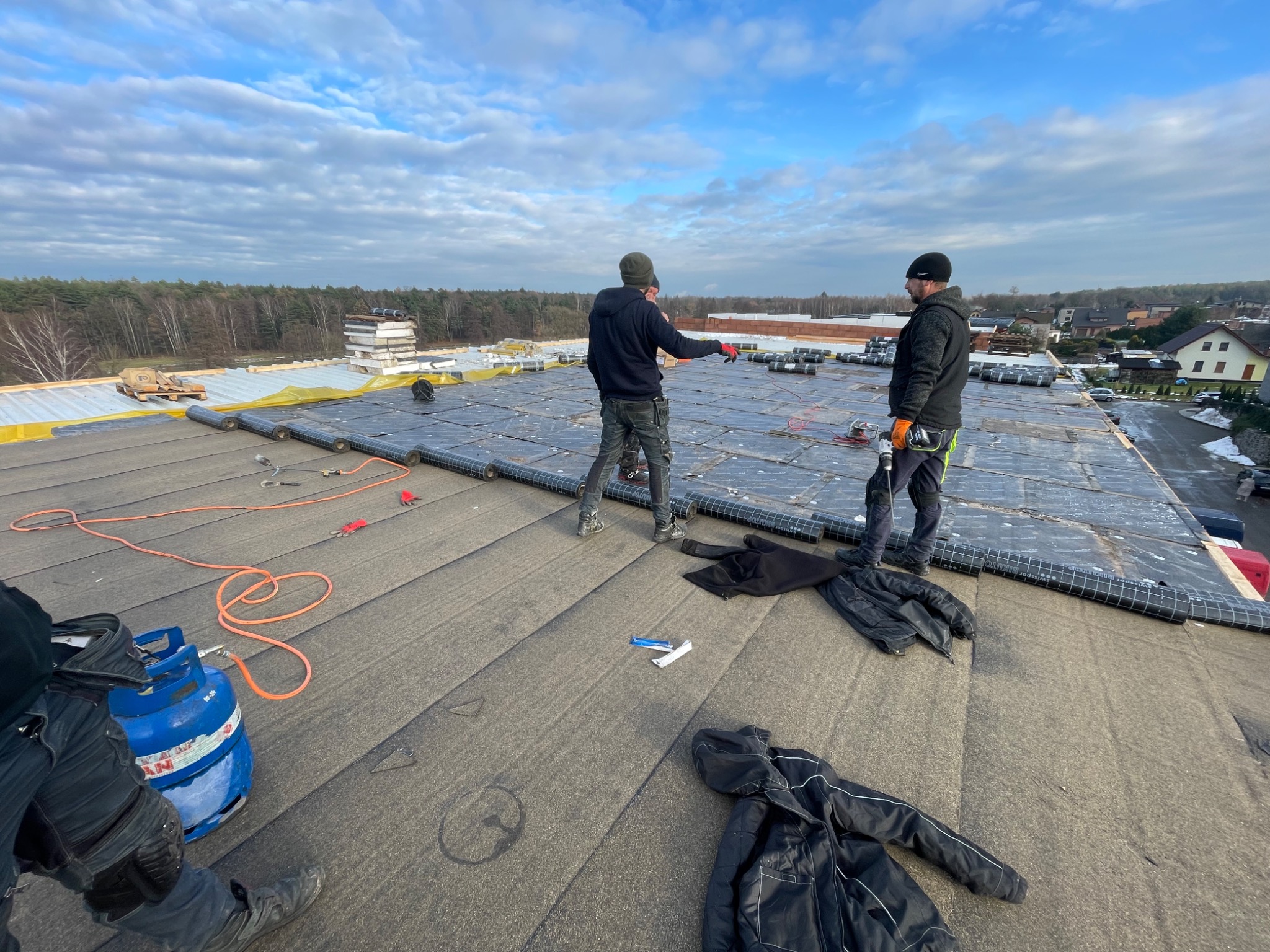 Dwóch dekarzy na dachu z papy termozgrzewalnej, jeden wskazujący na odległość, butla gazowa i narzędzia w tle, widok na las i zabudowania.