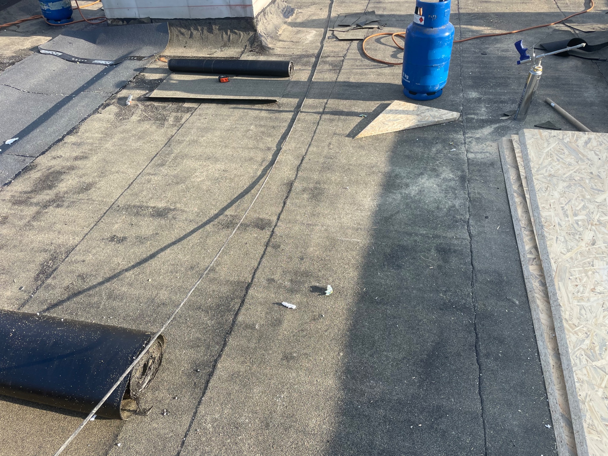 Widok z góry na płaski dach pokryty papą, z widocznymi rolkami papy, butlą gazową, deskami OSB i narzędziami podczas prac dekarskich.