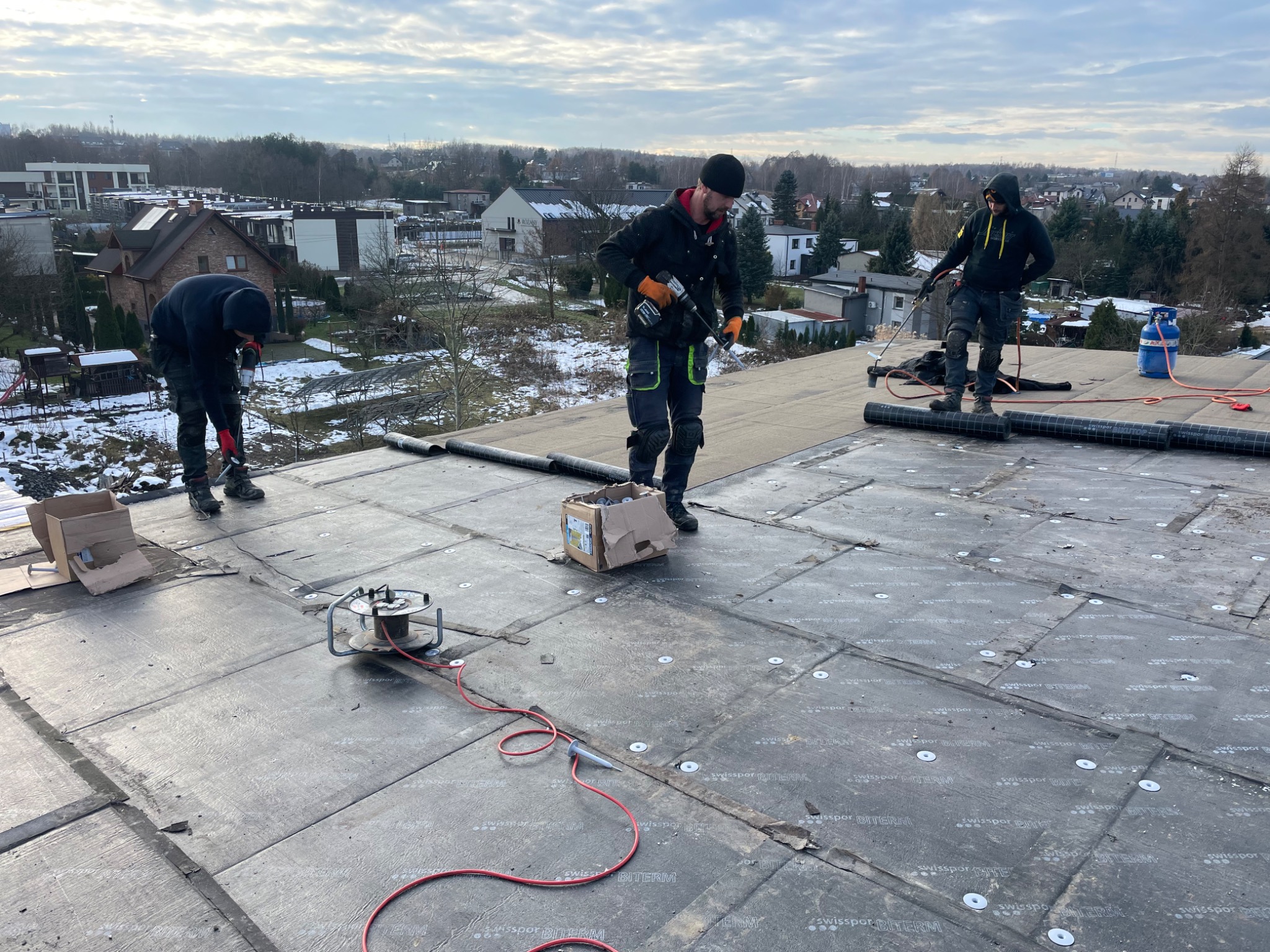 Trzech dekarzy pracuje na dachu pokrytym papą bitumiczną z widocznymi mocowaniami, w tle zabudowa miejska i drzewa, pochmurne niebo.