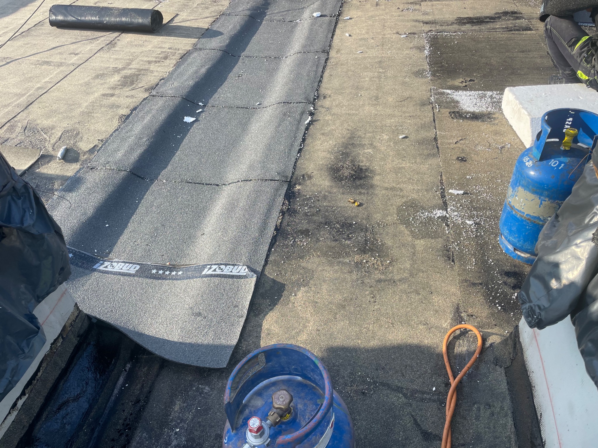 Fragment dachu płaskiego w trakcie prac z użyciem papy termozgrzewalnej marki Izobud, widoczne butle z gazem i rolka papy, z bliska.