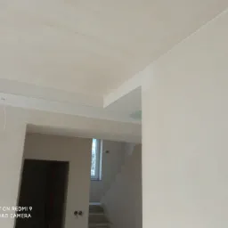 Wnętrze w trakcie remontu: sufit z płyt kartonowo-gipsowych, częściowo pomalowane ściany, widok na schody i okno w tle, niedokończone wykończenia.