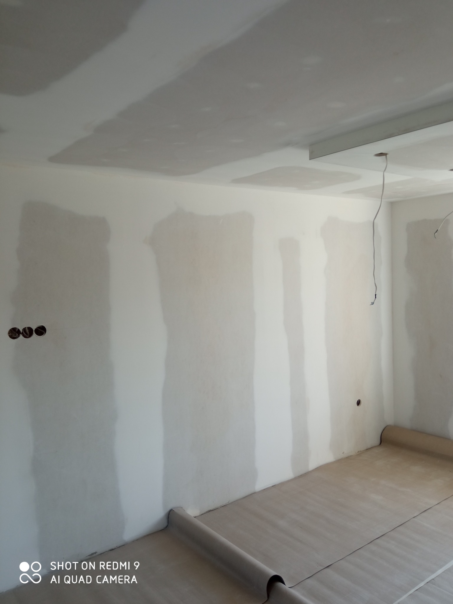 Pokój w trakcie prac wykończeniowych: ściany z widocznymi łączeniami płyt, częściowo wyszpachlowane, zabezpieczona podłoga, wystające przewody elektryczne.