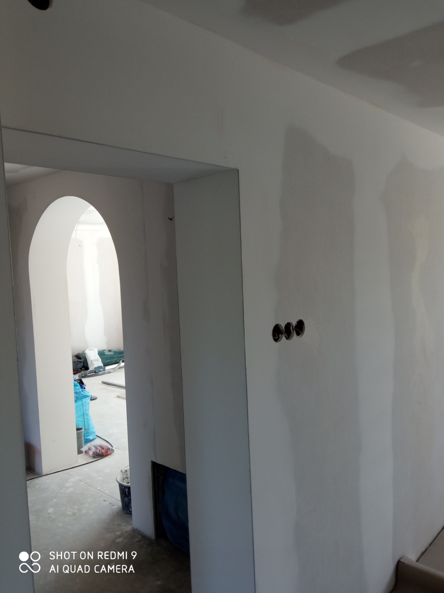 Wnętrze w trakcie prac wykończeniowych: ściany po szpachlowaniu, widoczne puszki elektryczne, przejście z łukiem, narzędzia i wiadro w tle.