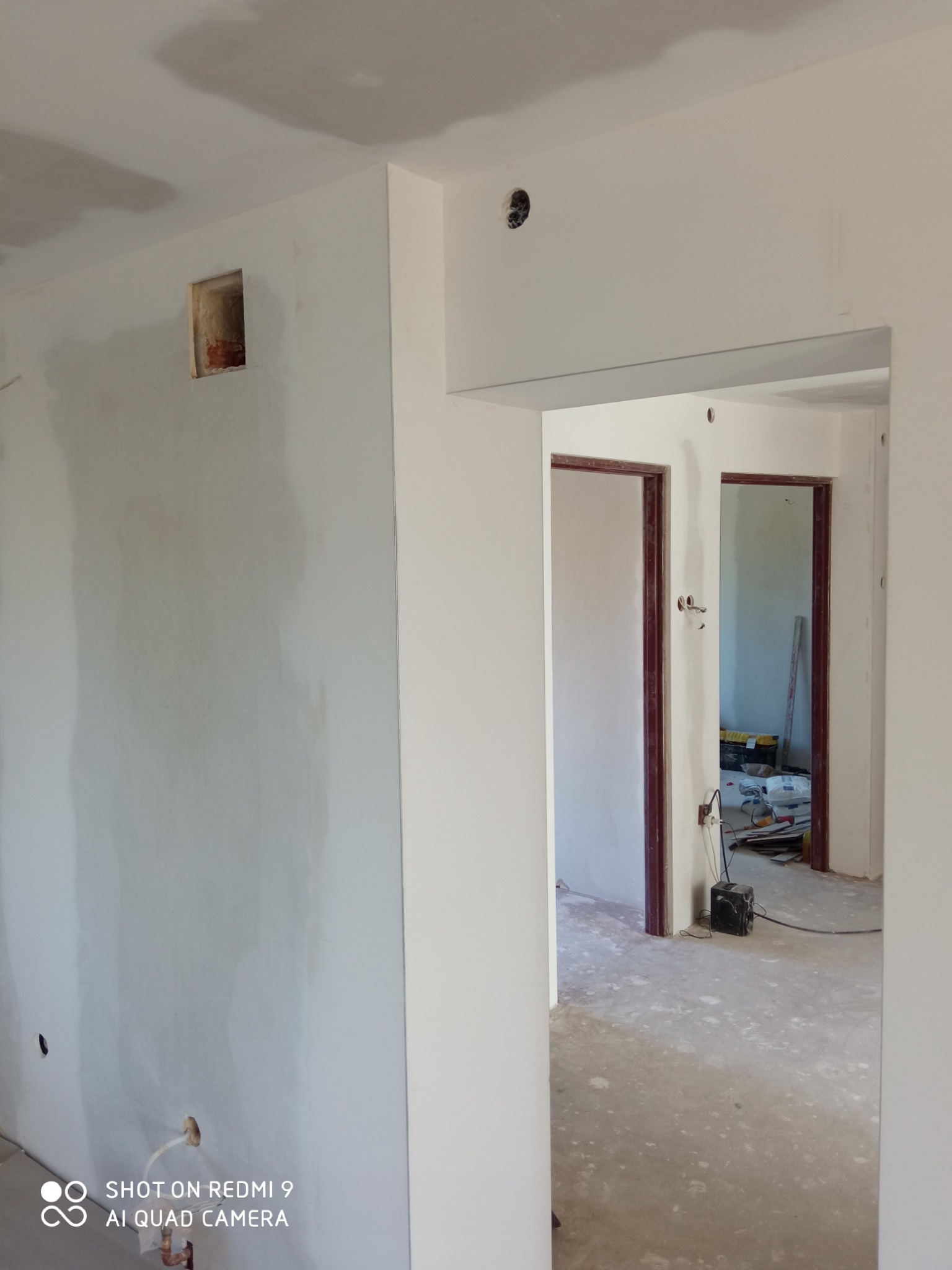 Wnętrze w trakcie prac wykończeniowych: ściany częściowo pokryte gładzią gipsową, widoczne otwory na instalacje elektryczne i hydrauliczne, surowy stan podłogi, ościeżnice drzwiowe bez skrzydeł.