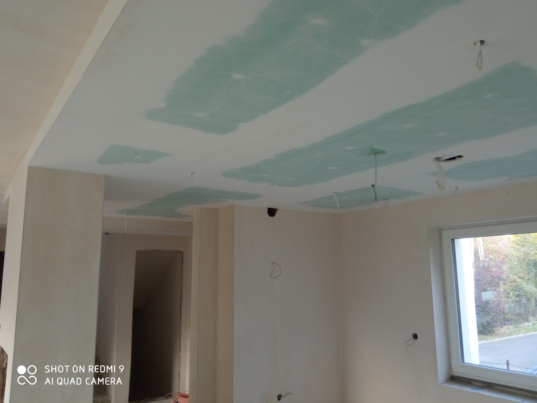 Wnętrze w trakcie prac wykończeniowych: sufit z widocznymi łączeniami płyt gipsowo-kartonowych przed szpachlowaniem, ściany przygotowane do malowania, widoczne przewody elektryczne, okno z widokiem...