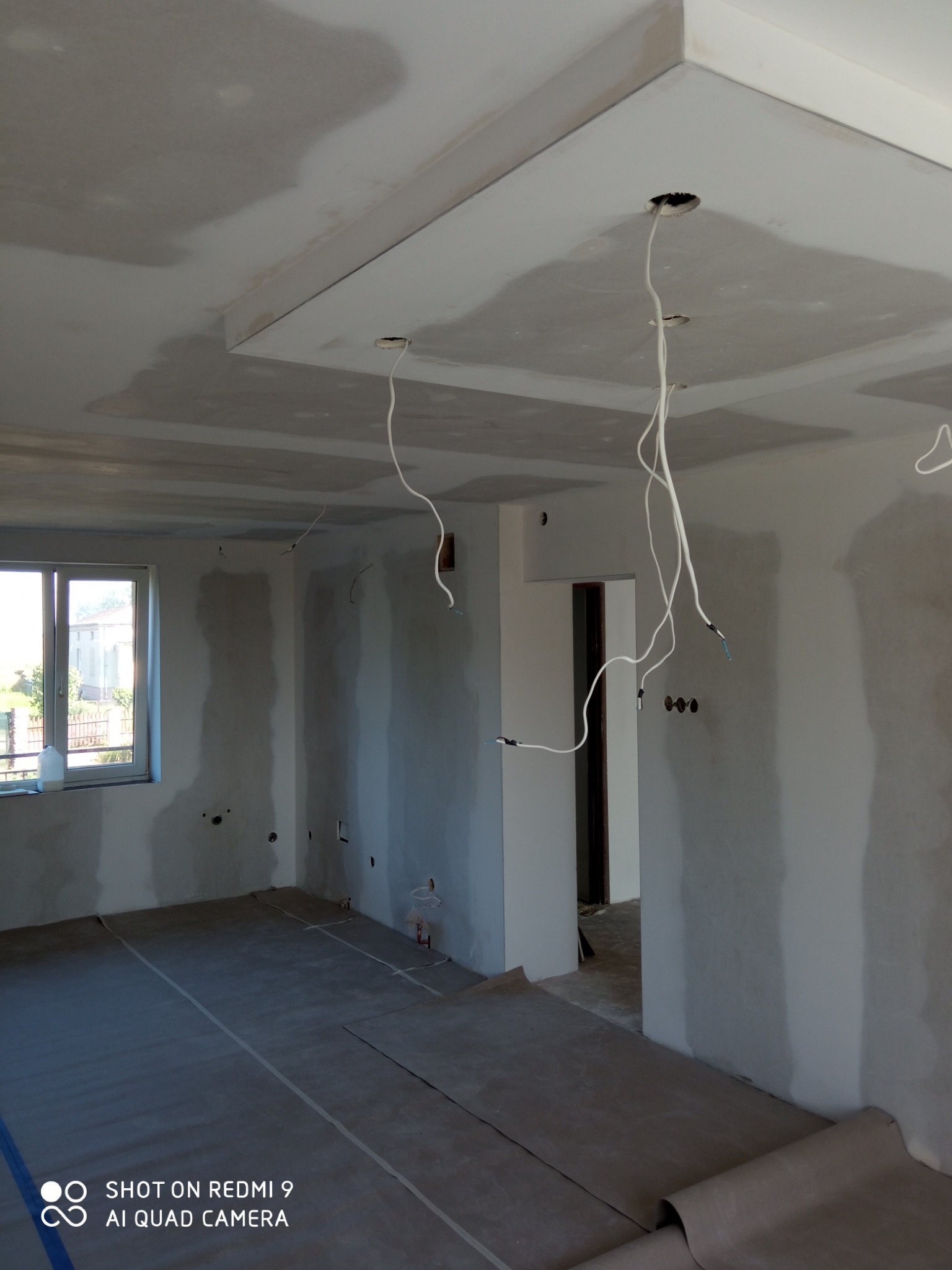 Wnętrze w trakcie prac wykończeniowych: surowa ściana, podłoga zabezpieczona folią, widoczne kable wystające z otworów w suficie podwieszanym z płyty gipsowej, okno z widokiem na zewnątrz.