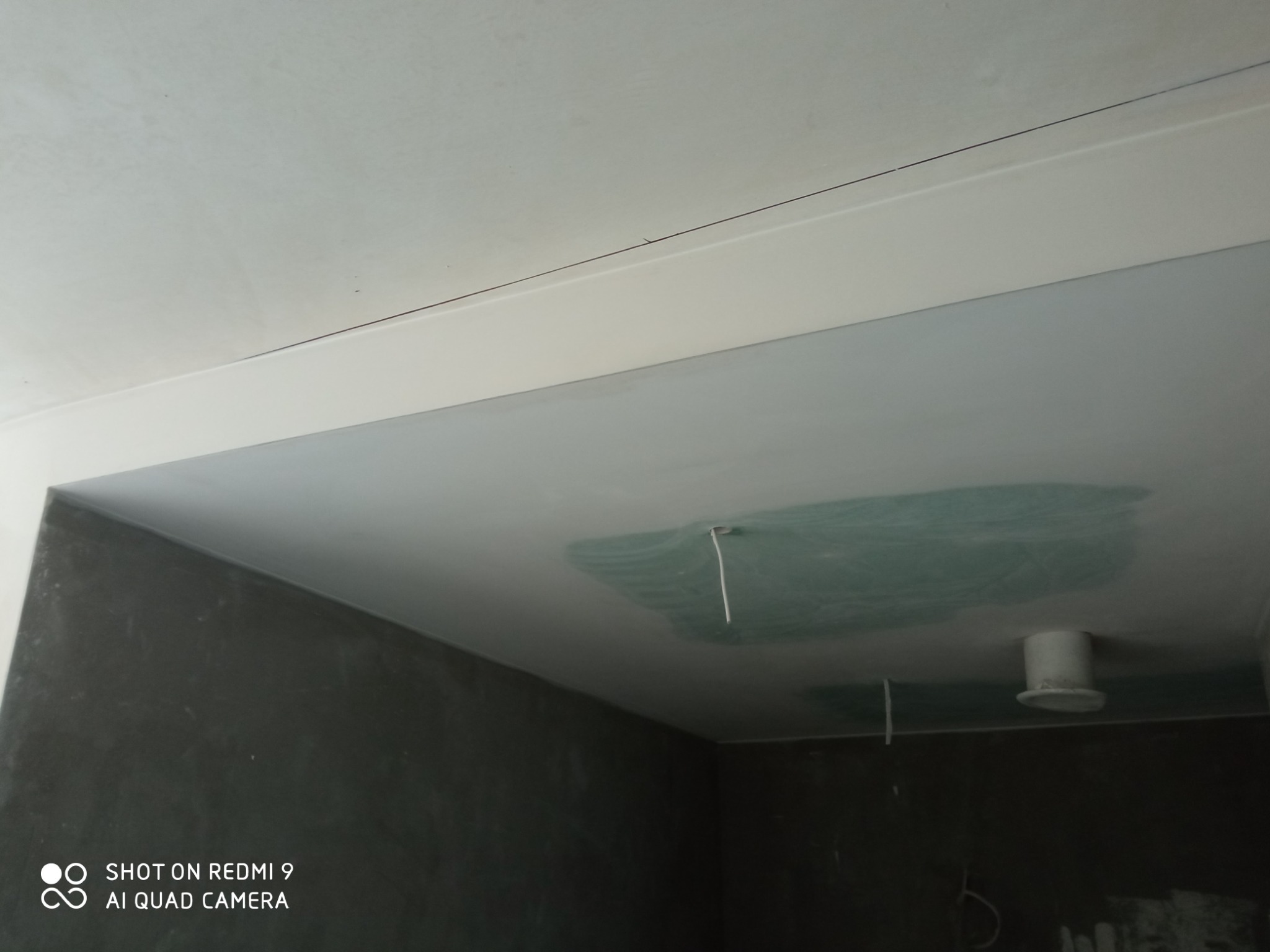 Niedokończony sufit z widocznym łączeniem płyt, wystającym kablem i rurą wentylacyjną, surowe ściany w tle.