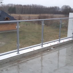 Stylbram - Szklana balustrada z szarymi słupkami na balkonie w trakcie montażu, widoczne elementy poziomowania płytek, w tle dom z drewnianą elewacją i zalesiony krajobraz.