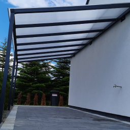 Stylbram - Nowoczesna, minimalistyczna pergola z czarną konstrukcją i przezroczystym zadaszeniem, przylegająca do białej ściany budynku z widocznym kranem na elewacji. Podłoże wyłożone szarą kostką brukową,...