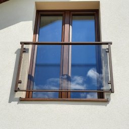 Stylbram - Elewacja budynku z dwoma oknami z brązowymi ramami. Górne okno posiada przeszkloną balustradę z brązowymi elementami mocującymi.