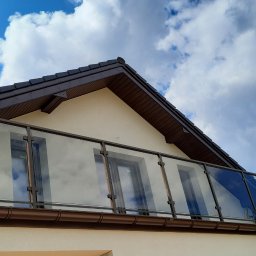 Stylbram - Szklana balustrada balkonowa z brązowymi ramami, widok z dołu na tle błękitnego nieba z chmurami, odbicie okien w szkle.