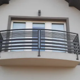 Czarna, metalowa balustrada balkonowa z poziomymi prętami, zamontowana na balkonie z trzema białymi oknami w szarym budynku, widok z dołu.