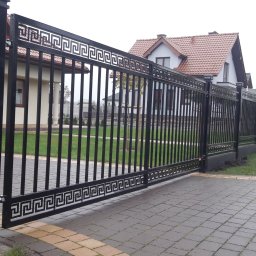 Stylbram - Automatyczna brama przesuwna z czarnych, pionowych paneli z motywem greckiego klucza, zamontowana na posesji w Siedliszcze, z widocznym domem w tle.