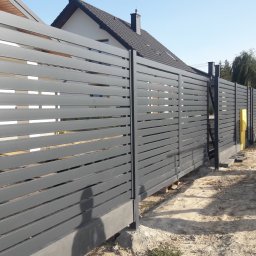 Stylbram - Nowoczesne, poziome, grafitowe przęsła ogrodzeniowe z metalu, zamontowane na betonowej podmurówce, widok z zewnątrz posesji, z żółtą skrzynką gazową w tle.