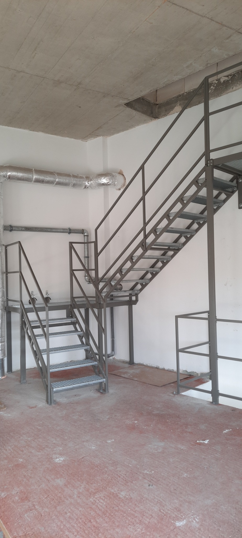 Metalowe schody przemysłowe z platformą, kratowe stopnie, szare rury wentylacyjne przy białej ścianie, surowy betonowy sufit i czerwona posadzka w surowym stanie.