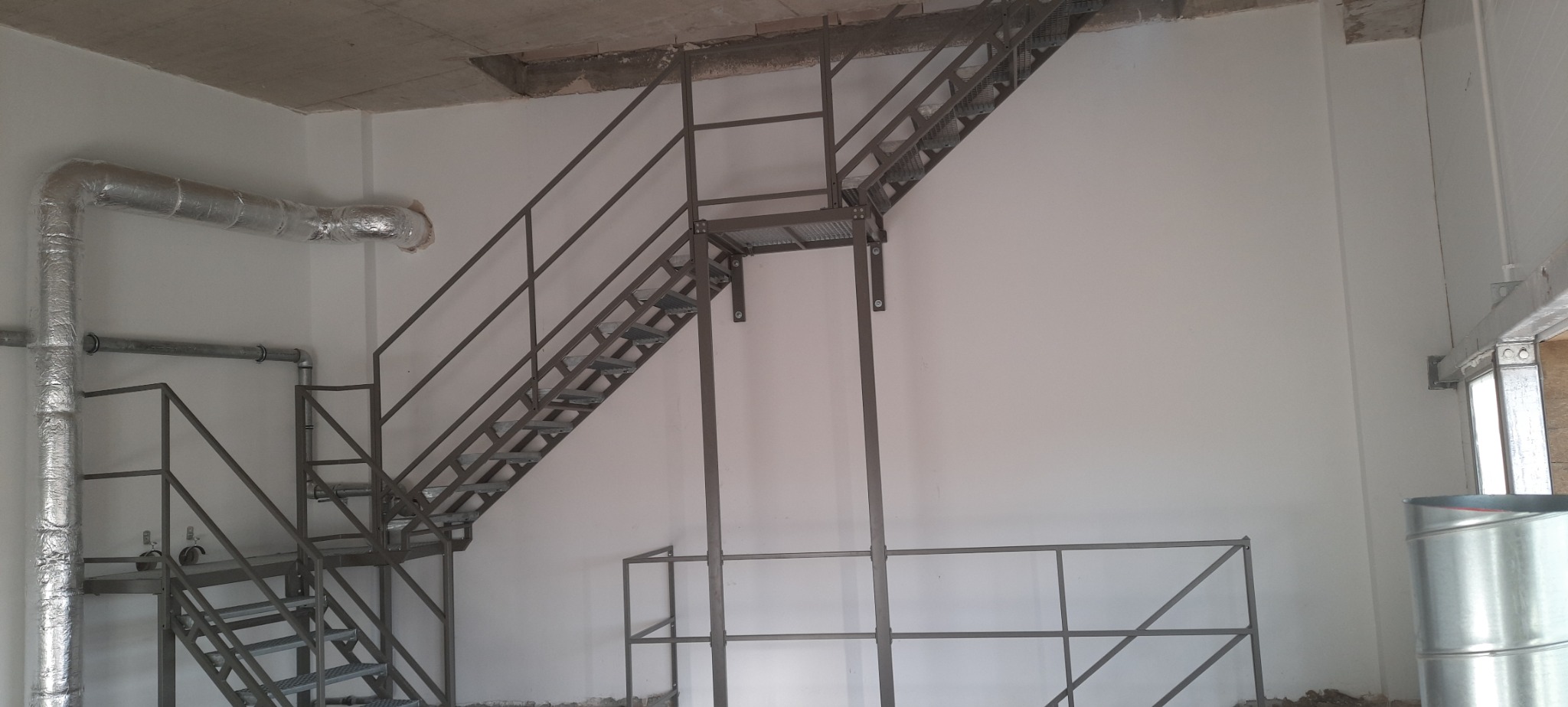Metalowe schody z platformą i poręczą, zamontowane wewnątrz budynku, widoczne rury wentylacyjne w izolacji.