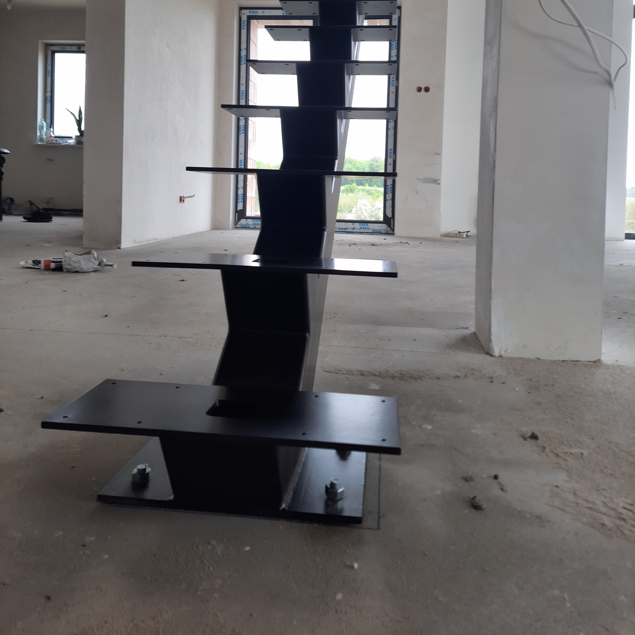 Surowe, czarne, metalowe schody bez poręczy, w trakcie montażu w nowym, surowym wnętrzu z widokiem na zieleń przez okno.