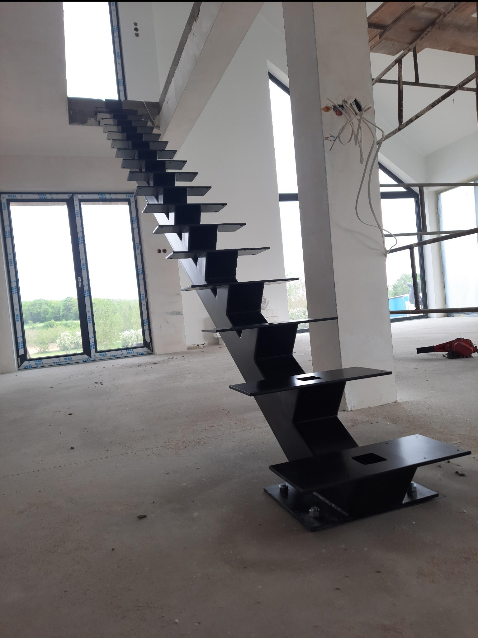 Nowoczesne, czarne schody metalowe z ażurową konstrukcją stopni, zamontowane w surowym wnętrzu budynku w trakcie budowy, widok z dołu na tle okien i nieotynkowanych ścian.