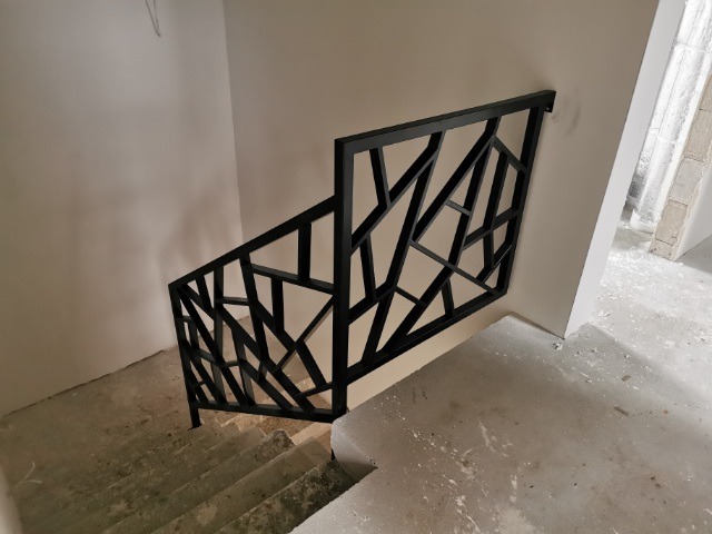 Czarna, metalowa balustrada schodowa o geometrycznym wzorze, zamontowana w surowym wnętrzu z betonowymi schodami i ścianami w trakcie wykończenia.