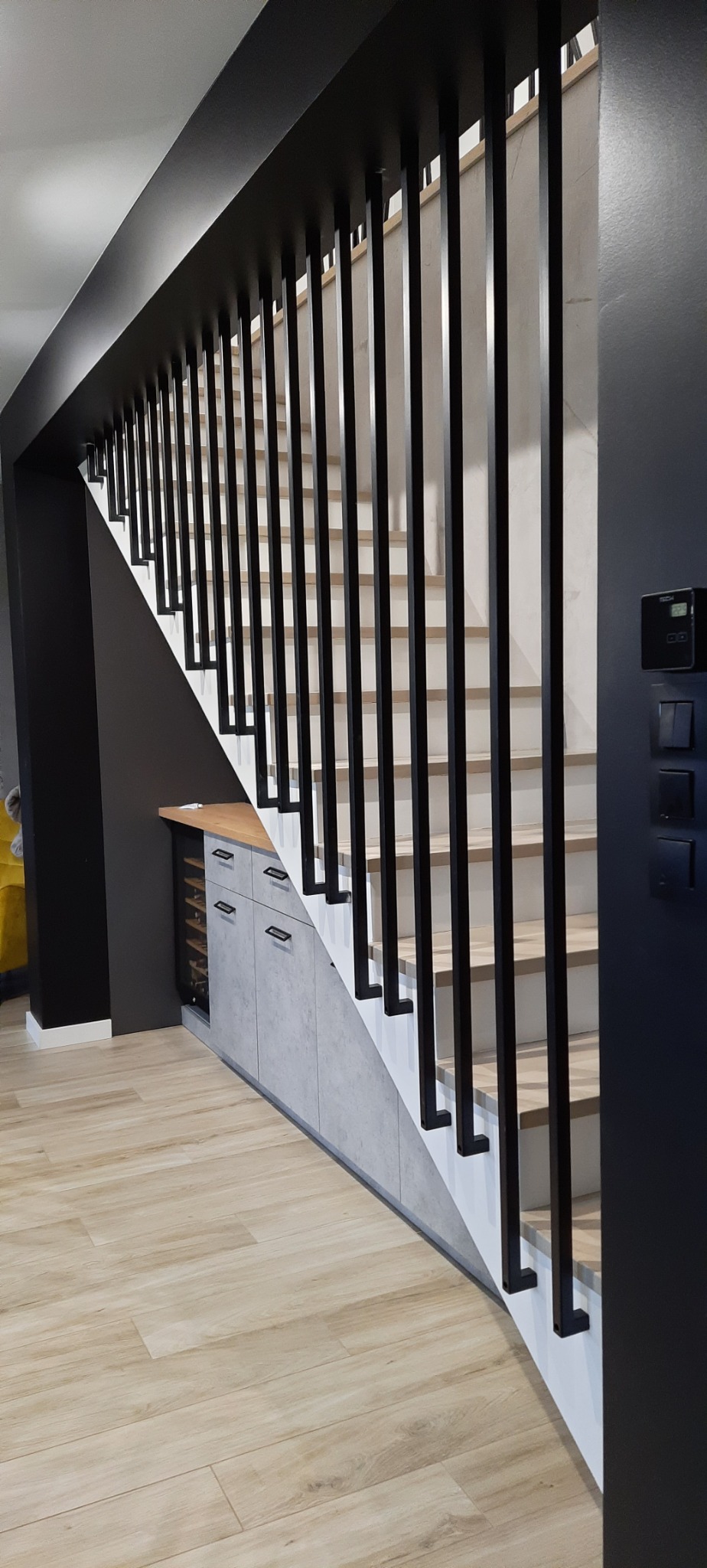 Nowoczesne schody wewnętrzne z drewnianymi stopniami i czarną metalową balustradą z pionowych słupków, zabudowa pod schodami z szufladami i szafkami wykończona w szarym betonie architektonicznym...