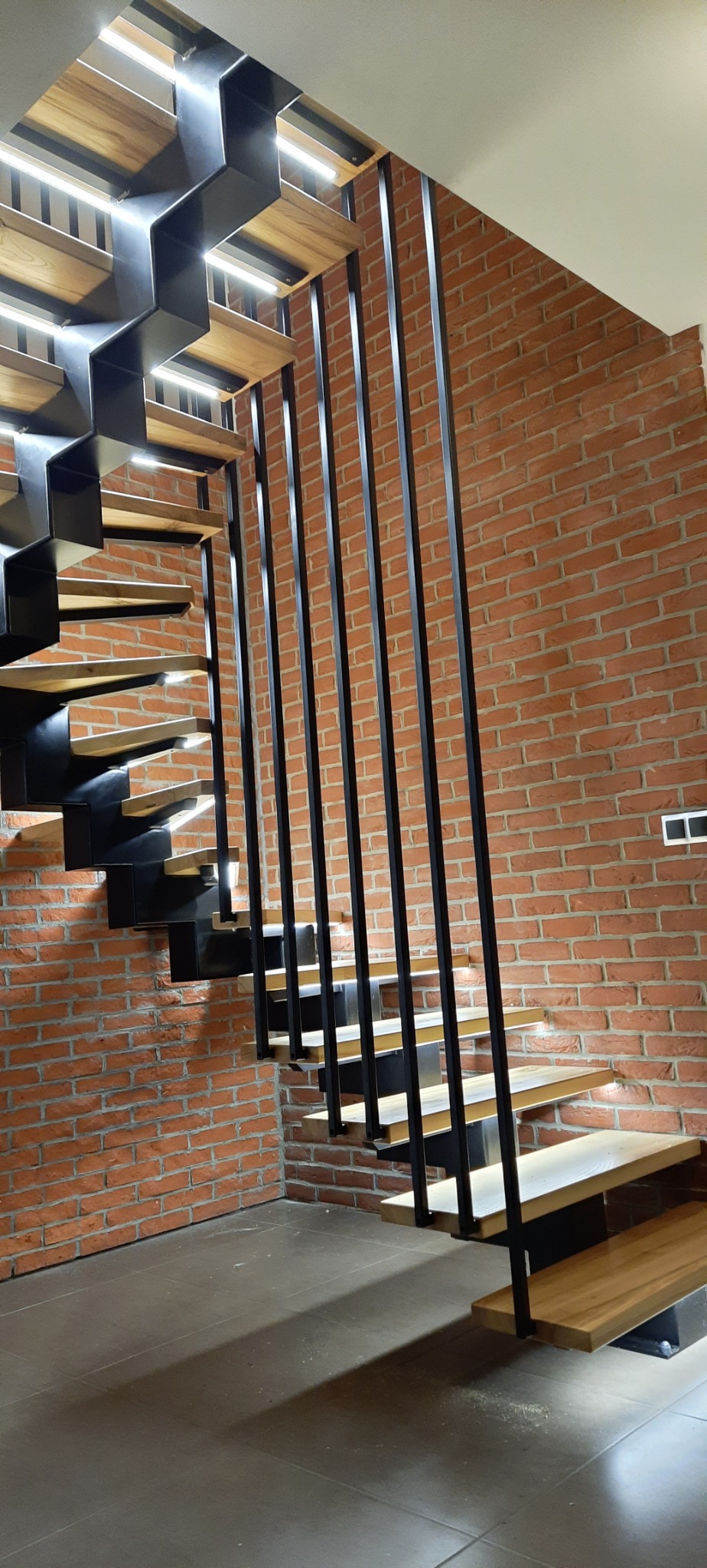 Nowoczesne schody metalowe z drewnianymi stopniami i minimalistyczną balustradą z czarnych prętów na tle ściany z czerwonej cegły.