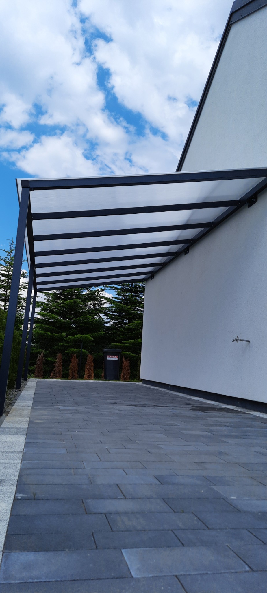 Nowoczesna, minimalistyczna pergola z czarną konstrukcją i przezroczystym zadaszeniem, przylegająca do białej ściany budynku z widocznym kranem na elewacji. Podłoże wyłożone szarą kostką brukową,...