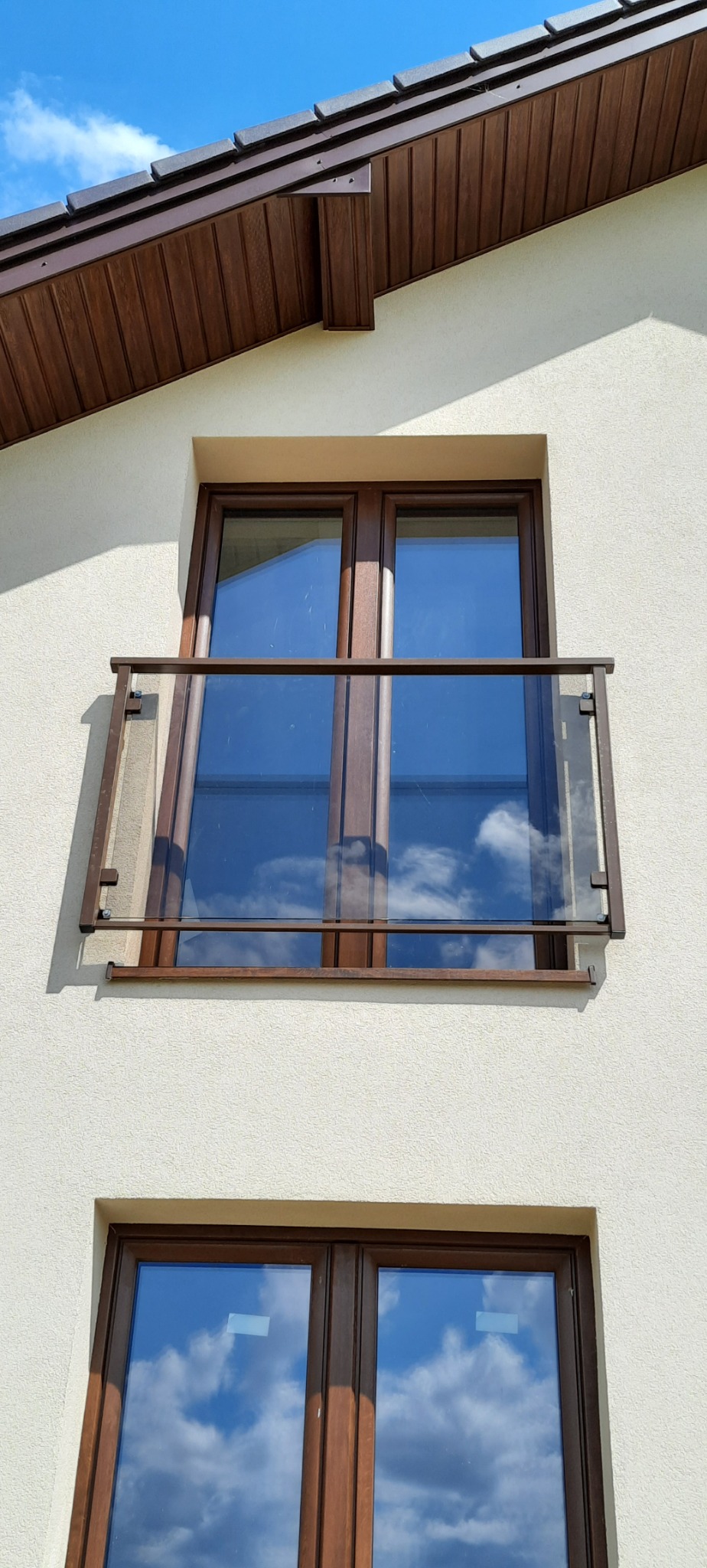 Elewacja budynku z dwoma oknami z brązowymi ramami. Górne okno posiada przeszkloną balustradę z brązowymi elementami mocującymi.