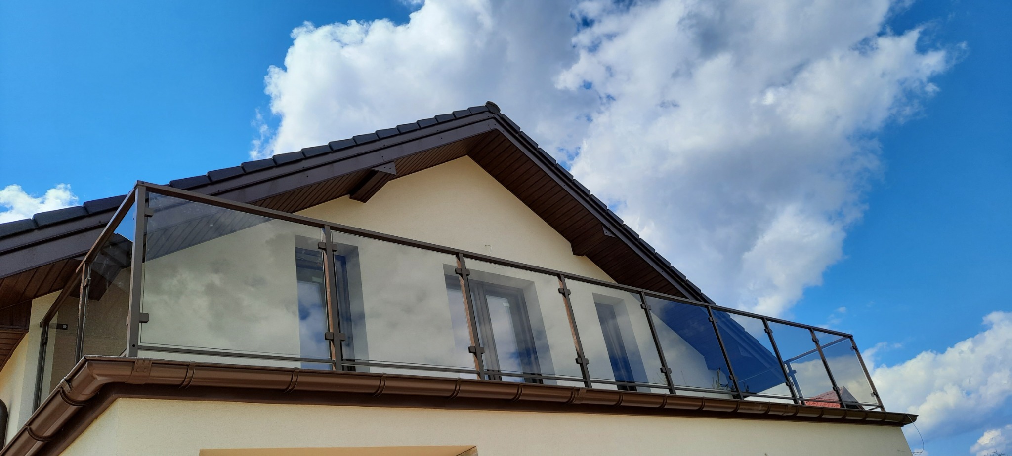 Szklana balustrada balkonowa z brązowymi ramami, widok z dołu na tle błękitnego nieba z chmurami, odbicie okien w szkle.