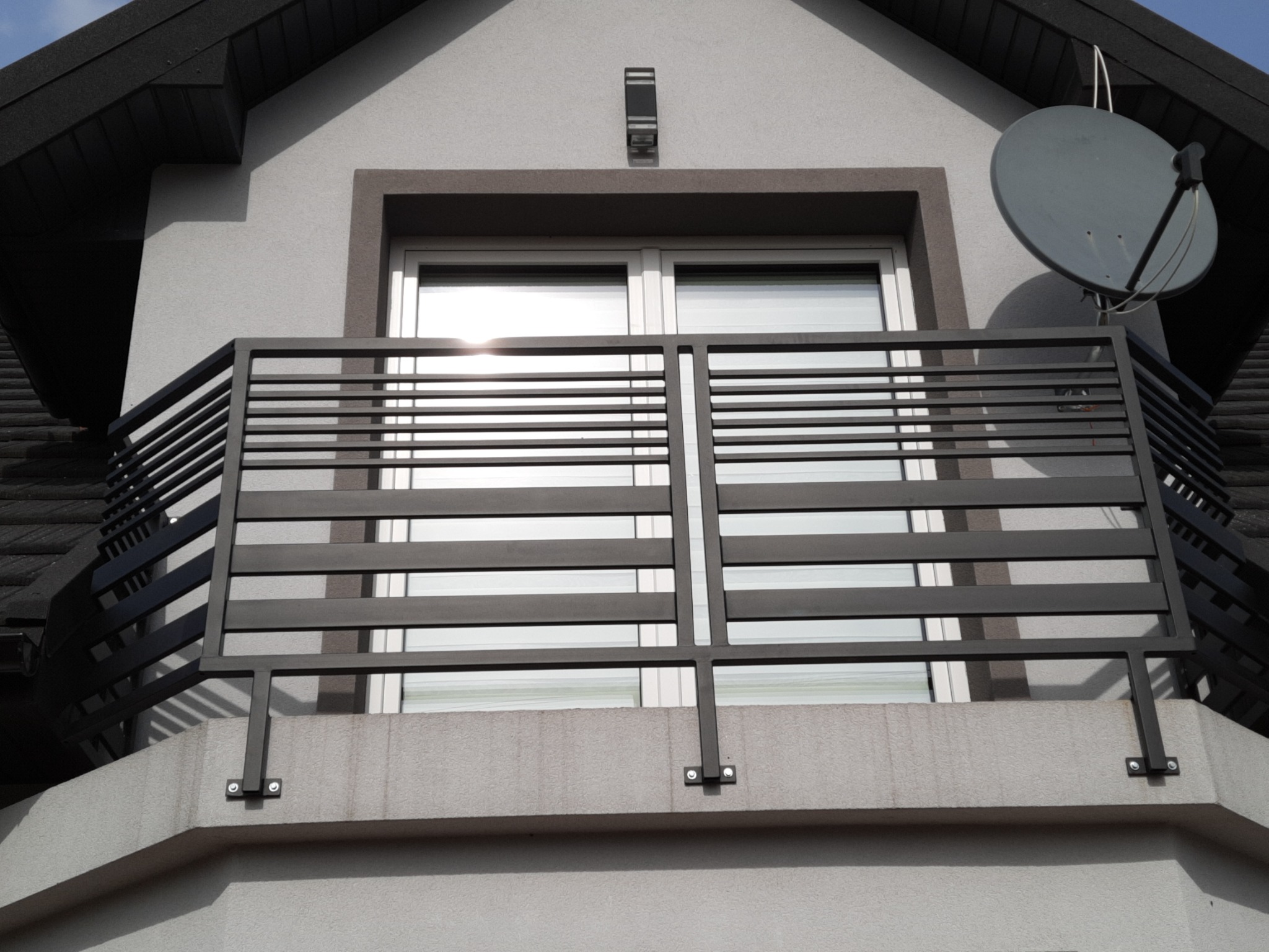 Szara balustrada balkonowa z poziomych profili, zamontowana na betonowej podstawie balkonu domu z szarą elewacją, widoczny fragment dachu i antena satelitarna.