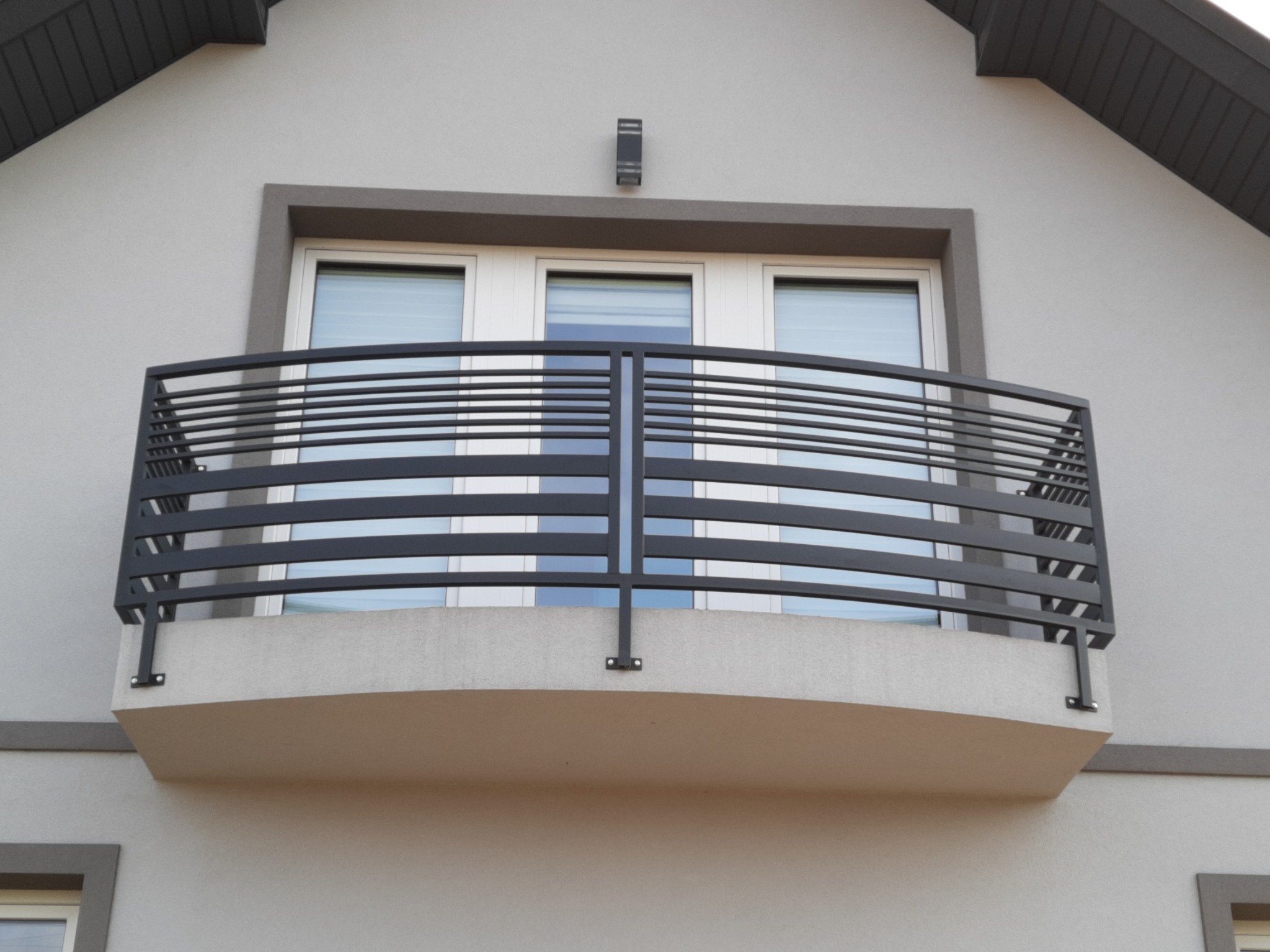 Czarna, metalowa balustrada balkonowa z poziomymi prętami, zamontowana na balkonie z trzema białymi oknami w szarym budynku, widok z dołu.