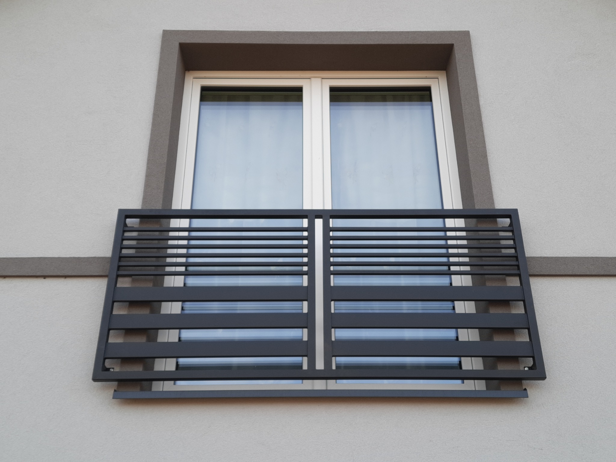 Nowoczesna, pozioma balustrada metalowa w kolorze antracytowym zamontowana na oknie budynku z szarą elewacją i białymi ramami okiennymi.