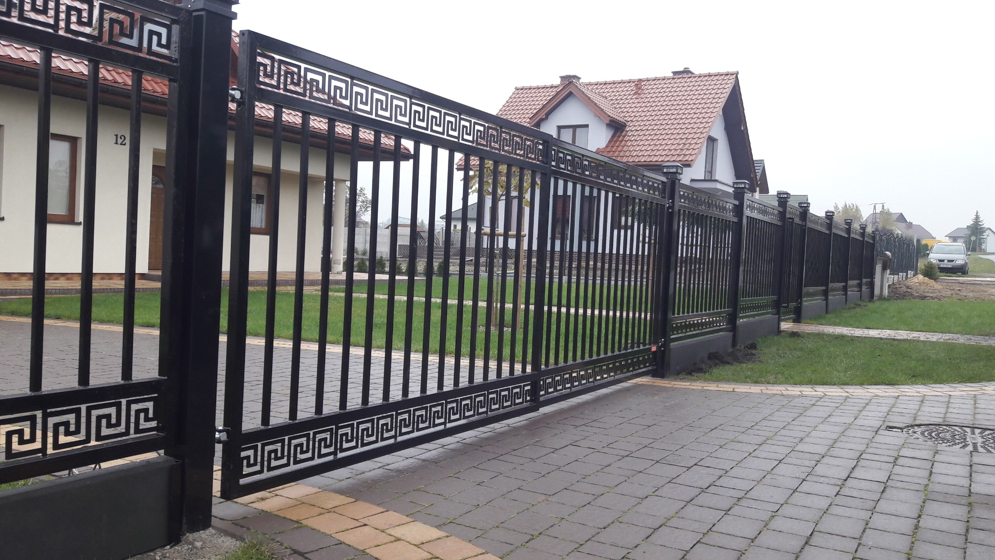 Automatyczna brama przesuwna z czarnych, pionowych paneli z motywem greckiego klucza, zamontowana na posesji w Siedliszcze, z widocznym domem w tle.