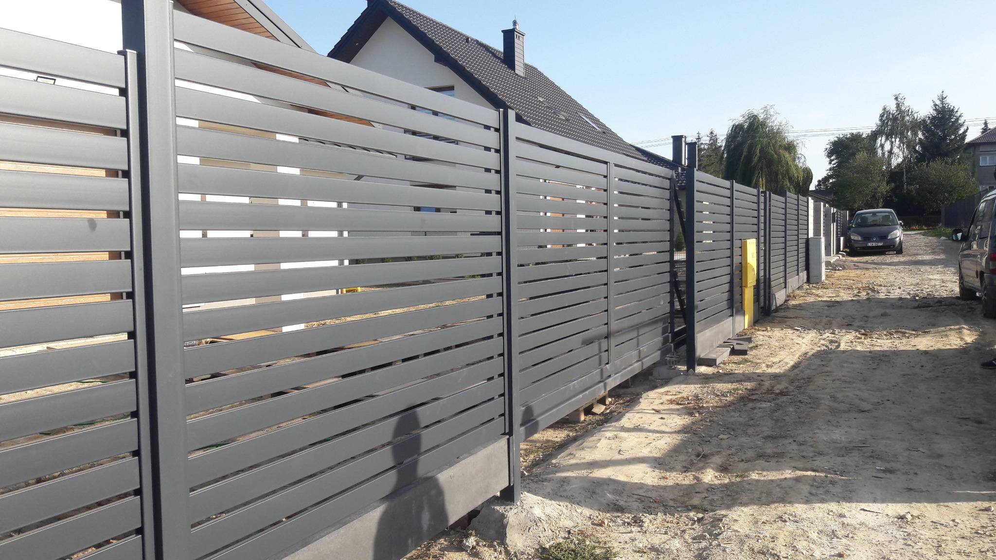 Nowoczesne, poziome, grafitowe przęsła ogrodzeniowe z metalu, zamontowane na betonowej podmurówce, widok z zewnątrz posesji, z żółtą skrzynką gazową w tle.