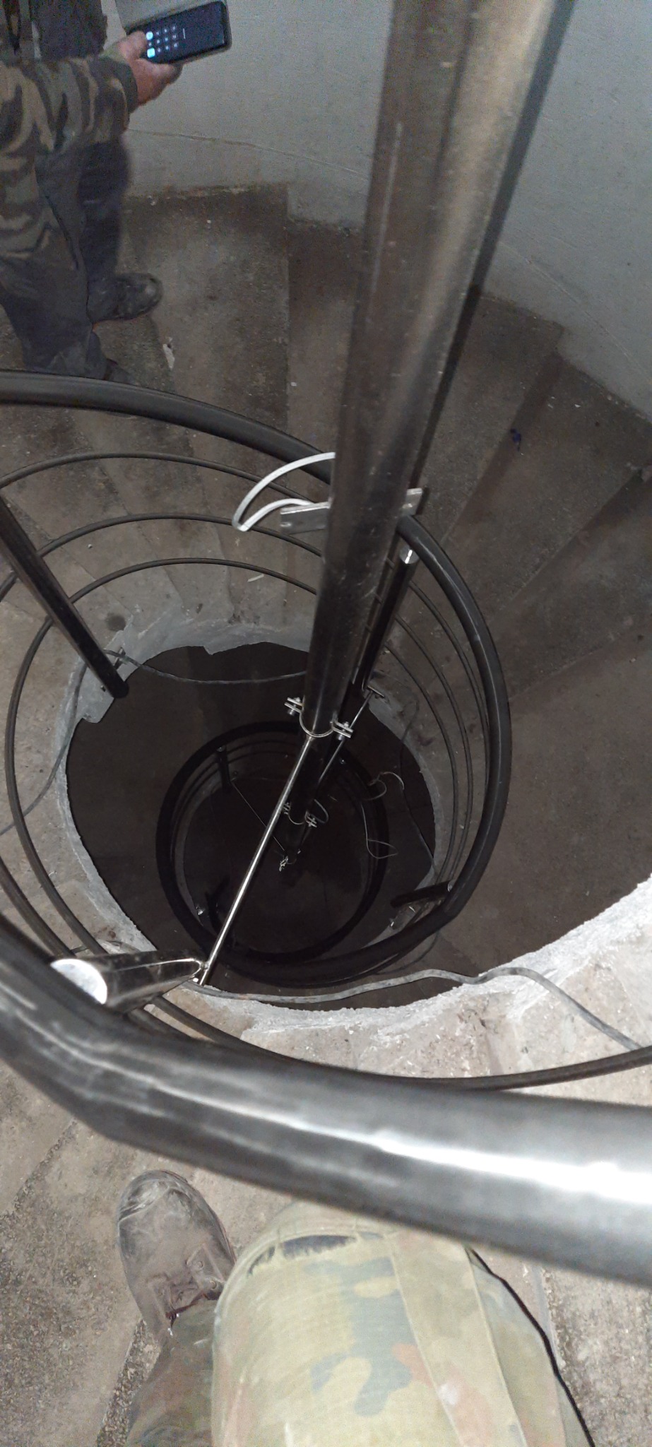 Wewnętrzny widok spiralnych schodów metalowych w surowym stanie, z widocznymi elementami montażowymi i fragmentem osoby w ubraniu roboczym.