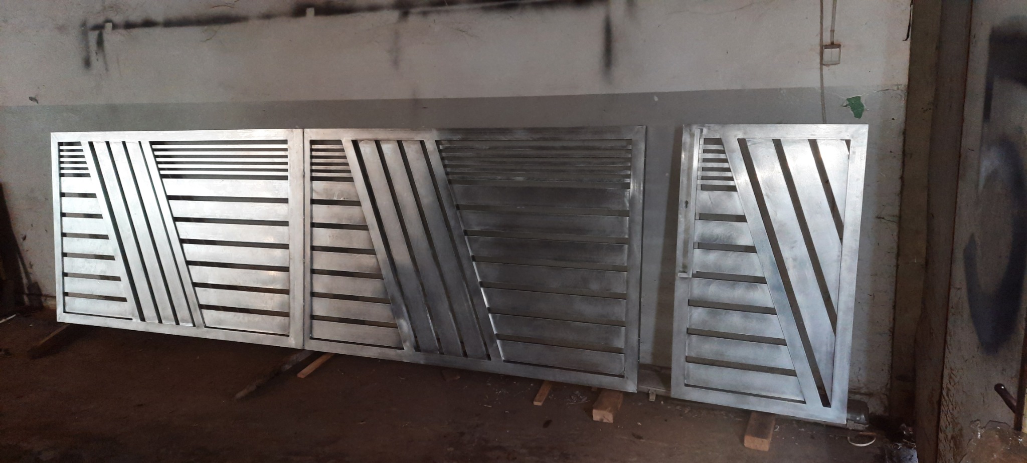 Trzy segmenty metalowej bramy z poziomymi i ukośnymi prętami, surowe wykończenie, ustawione w warsztacie na drewnianych klockach, ściany z zaciekami farby w tle.