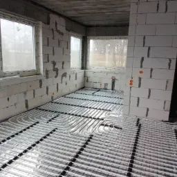 Białe rury ogrzewania podłogowego ułożone na czarnej siatce w surowym wnętrzu budynku z oknami i ścianami z betonu komórkowego.