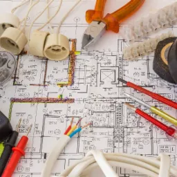 Plan instalacji elektrycznej z przyrządami i elementami: multimetr, przewody, puszki oświetleniowe, taśma izolacyjna i narzędzia na tle schematu.