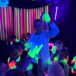 Ani - mani - Animator w białym fartuchu prezentuje eksperyment z dymem dla grupy dzieci w ciemnym pomieszczeniu z neonowymi dekoracjami i dyskotekową kulą.