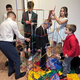Ani - mani - Grupa dzieci w strojach galowych buduje konstrukcję z kolorowych rurek na tle jasnej ściany. Zabawa edukacyjna podczas imprezy.