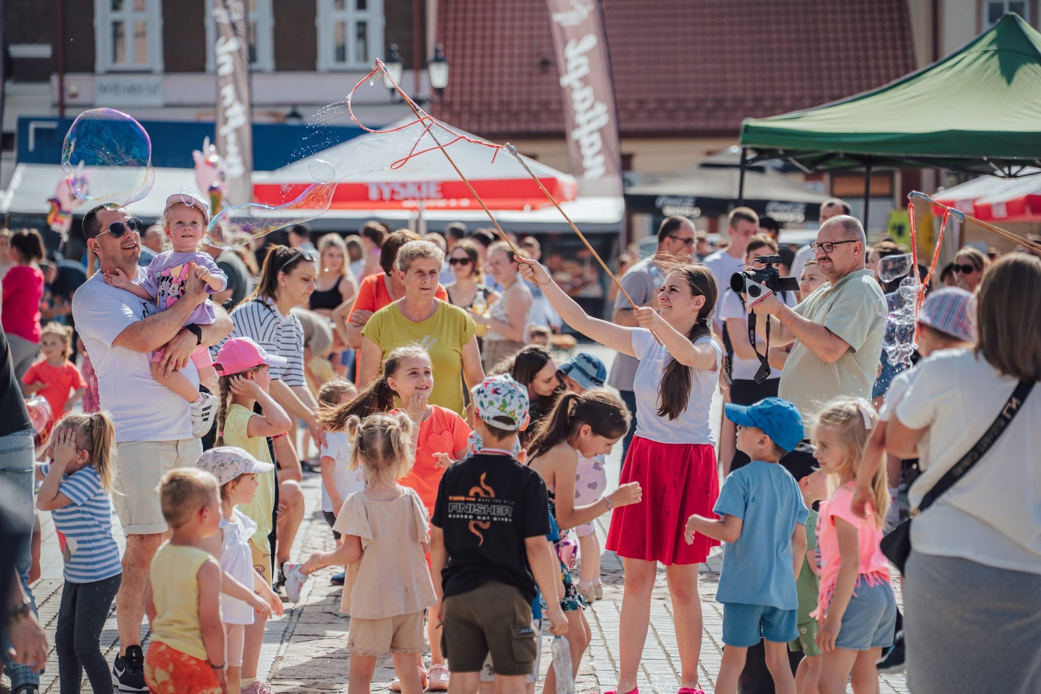 Animator robiący bańki mydlane na plenerowym evencie dla dzieci w słoneczny dzień. W tle tłum ludzi i budynki miejskie.