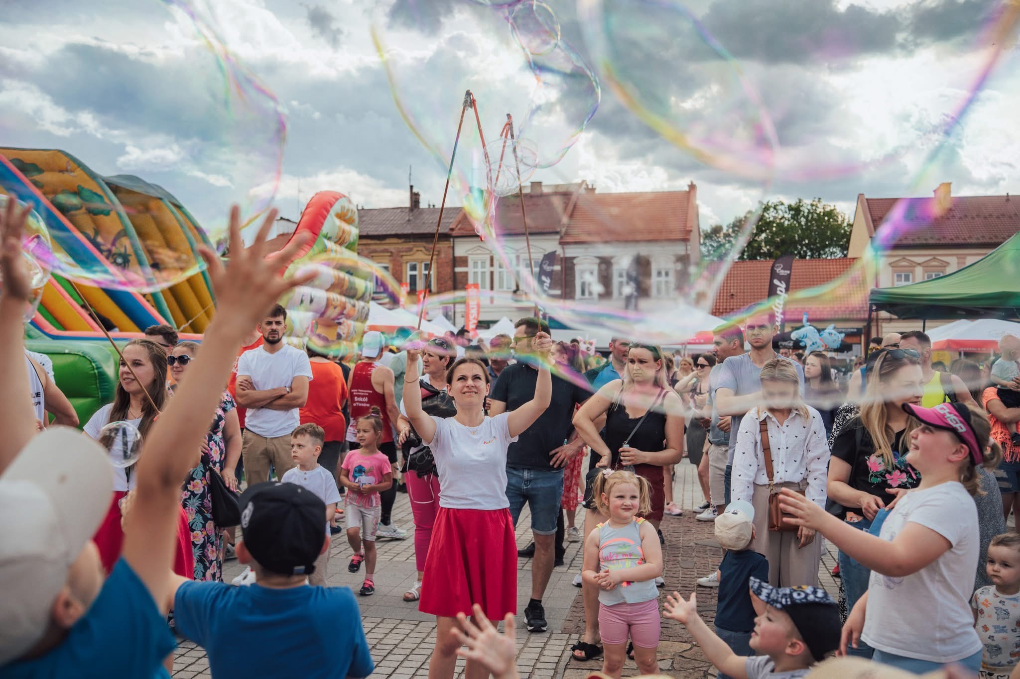 Animatorka w czerwonej spódnicy puszcza bańki mydlane na imprezie plenerowej, dzieci wyciągają ręce, w tle dmuchany zamek i tłum ludzi.