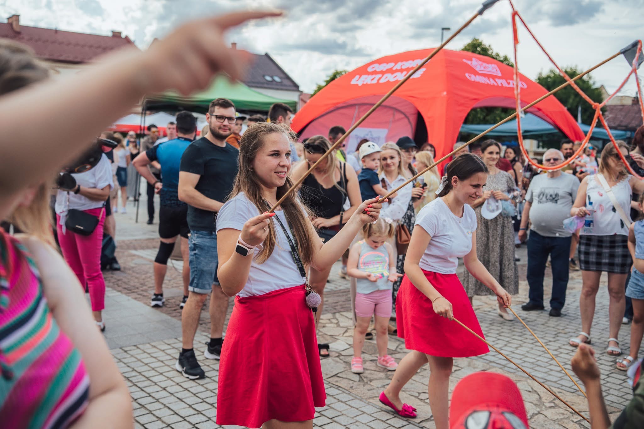 Animacje dla dzieci w Tarnowie: dziewczyny w czerwonych spódnicach prowadzą zabawę z kijkami na tle plenerowej imprezy z czerwonym namiotem w tle.