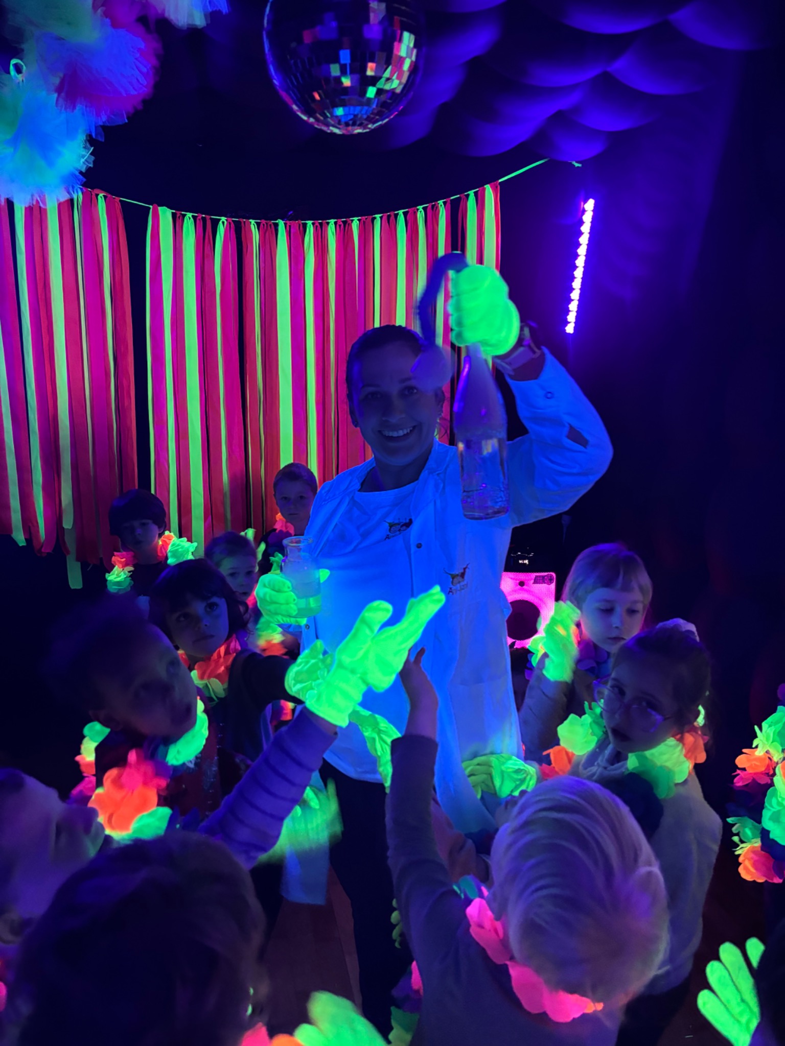 Animator w białym fartuchu prezentuje eksperyment z dymem dla grupy dzieci w ciemnym pomieszczeniu z neonowymi dekoracjami i dyskotekową kulą.