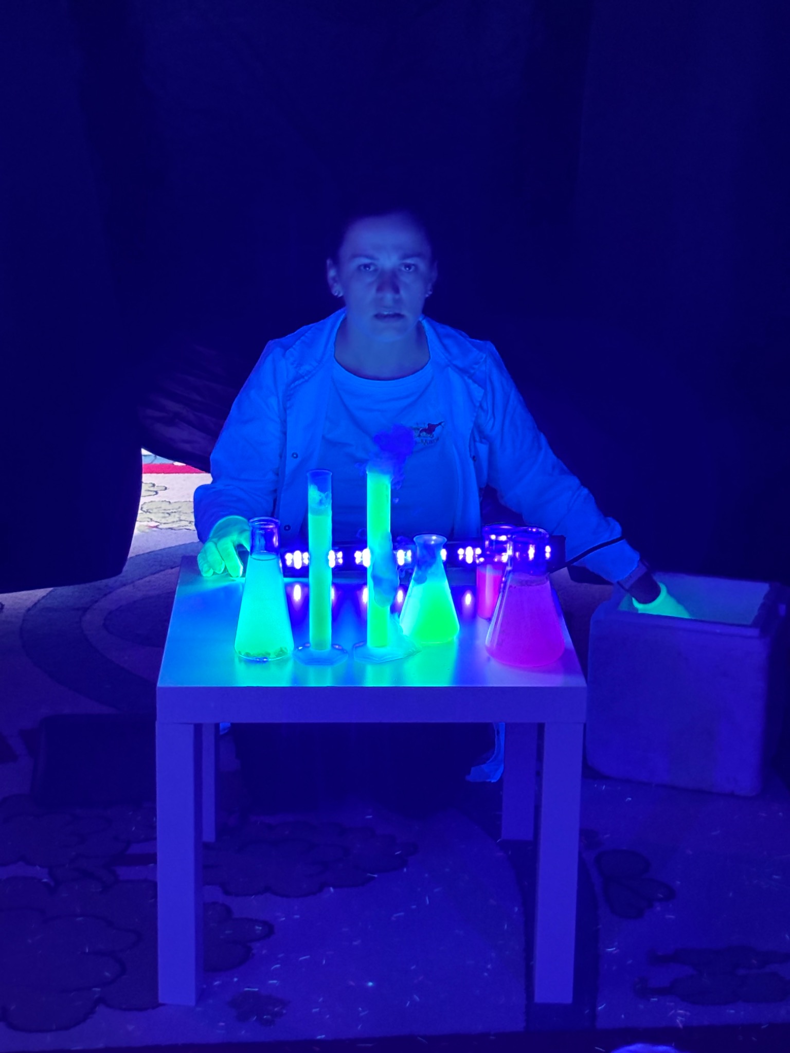 Osoba w białym fartuchu przeprowadza eksperymenty z fluorescencyjnymi płynami w probówkach i kolbach, oświetlonych światłem UV na stole. Mroczna sceneria.