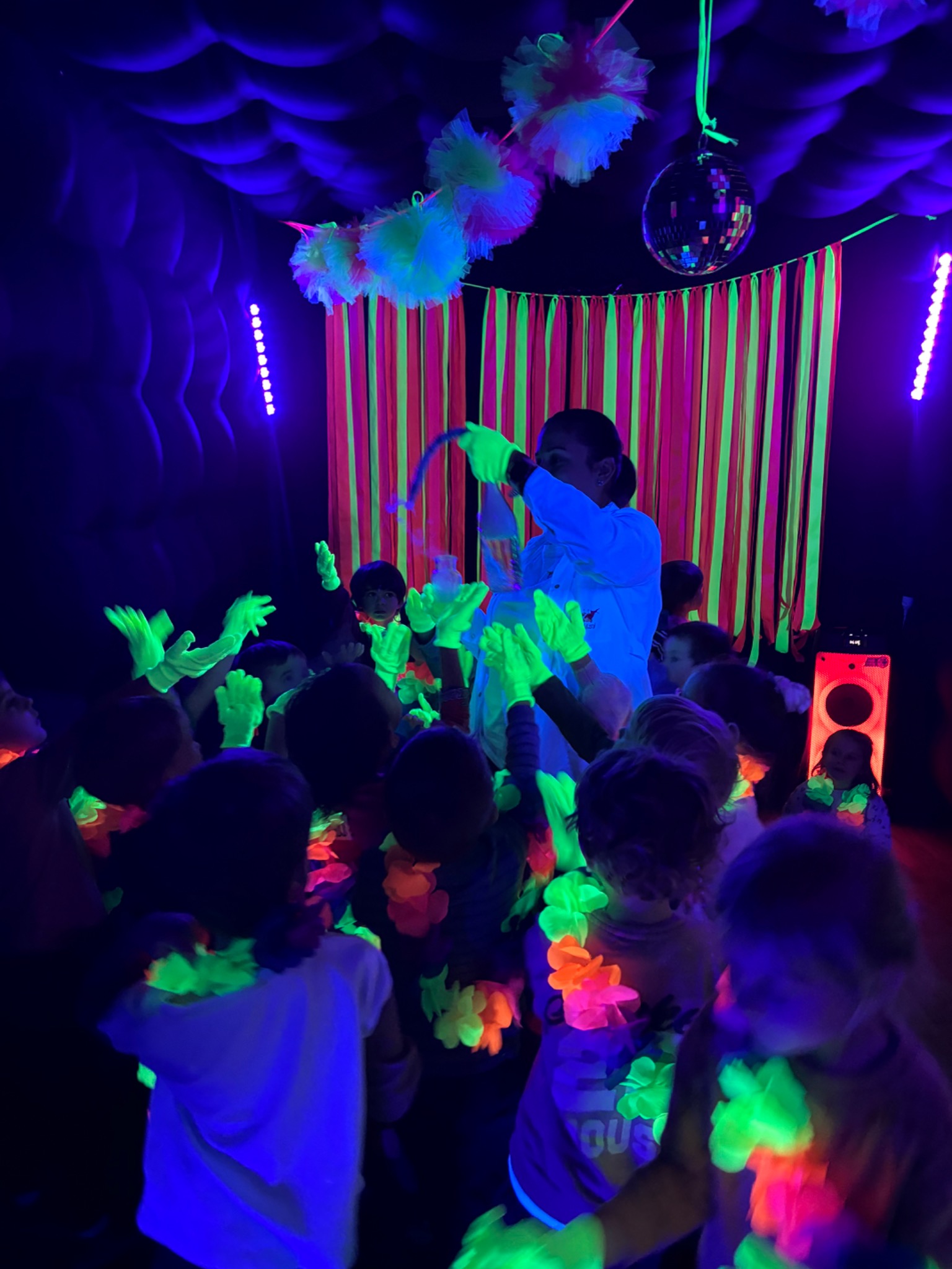 Animator w białym stroju prowadzi zabawę z dziećmi w ciemnym pomieszczeniu z dekoracjami UV, w tle kolumna głośnikowa i dyskotekowa kula. Dzieci mają na sobie świecące hawajskie naszyjniki.